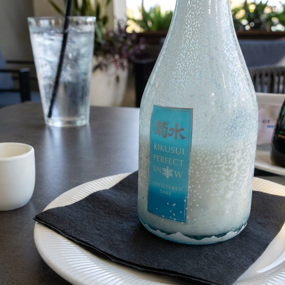 Table setting with a chilled Kikusui Perfect Snow unfiltered sake bottle, sake cup, water glass, and soy sauce.