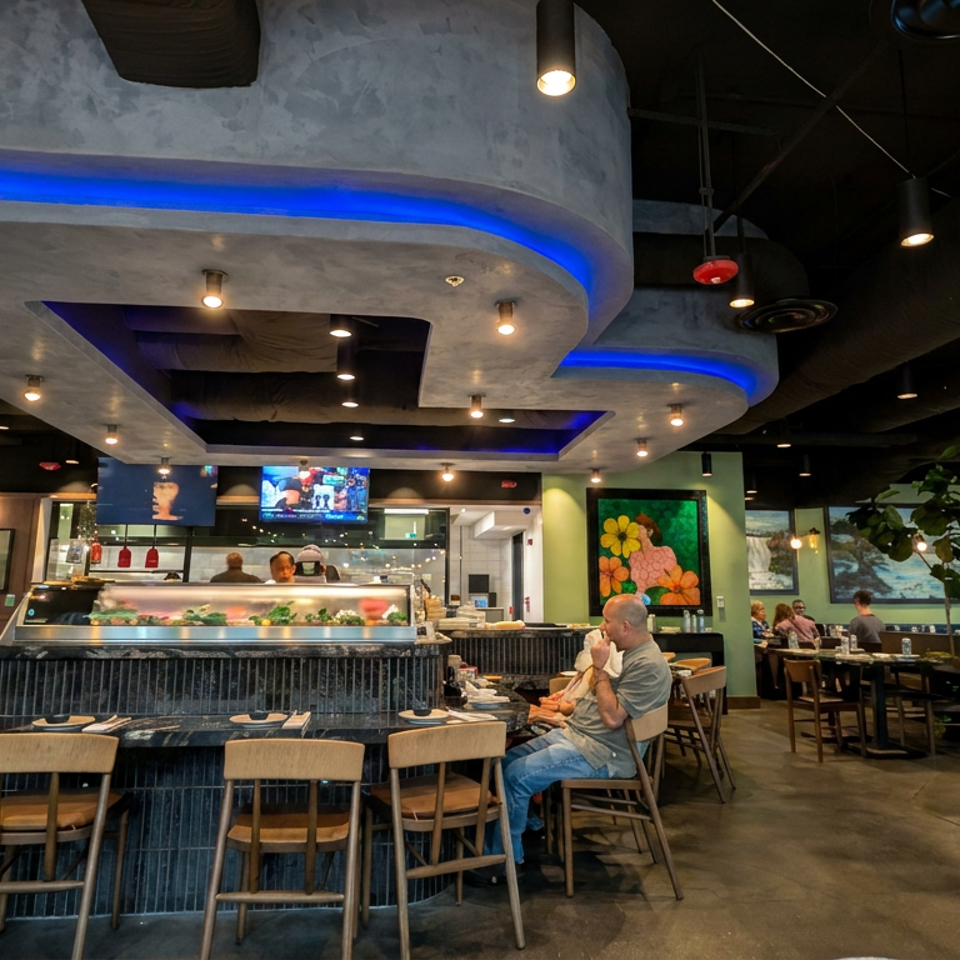 Spacious modern restaurant dining room with sushi counter, seated diners, sports on televisions, colorful artwork, and blue accent lighting.