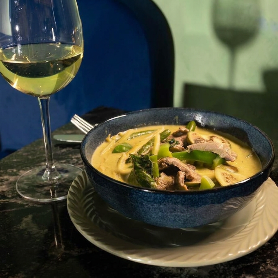 Blue bowl of creamy curry with vegetables and meat beside a glass of white wine on a dark restaurant table.
