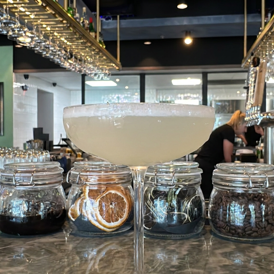 Cocktail glass filled with a creamy drink sits on a café counter beside jars of coffee beans, dried orange slices, and berries.
