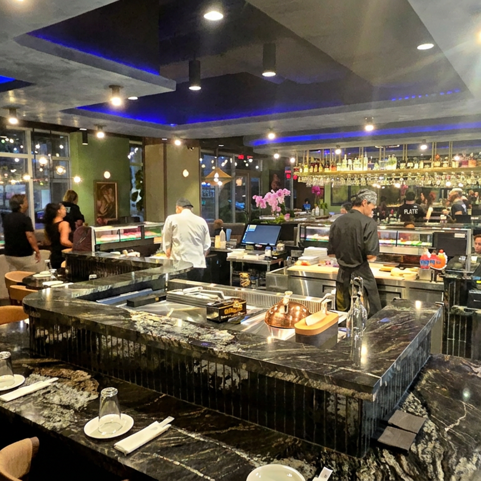 Modern restaurant interior with marble sushi bar, chefs preparing food, seated tables, and blue accent lighting.